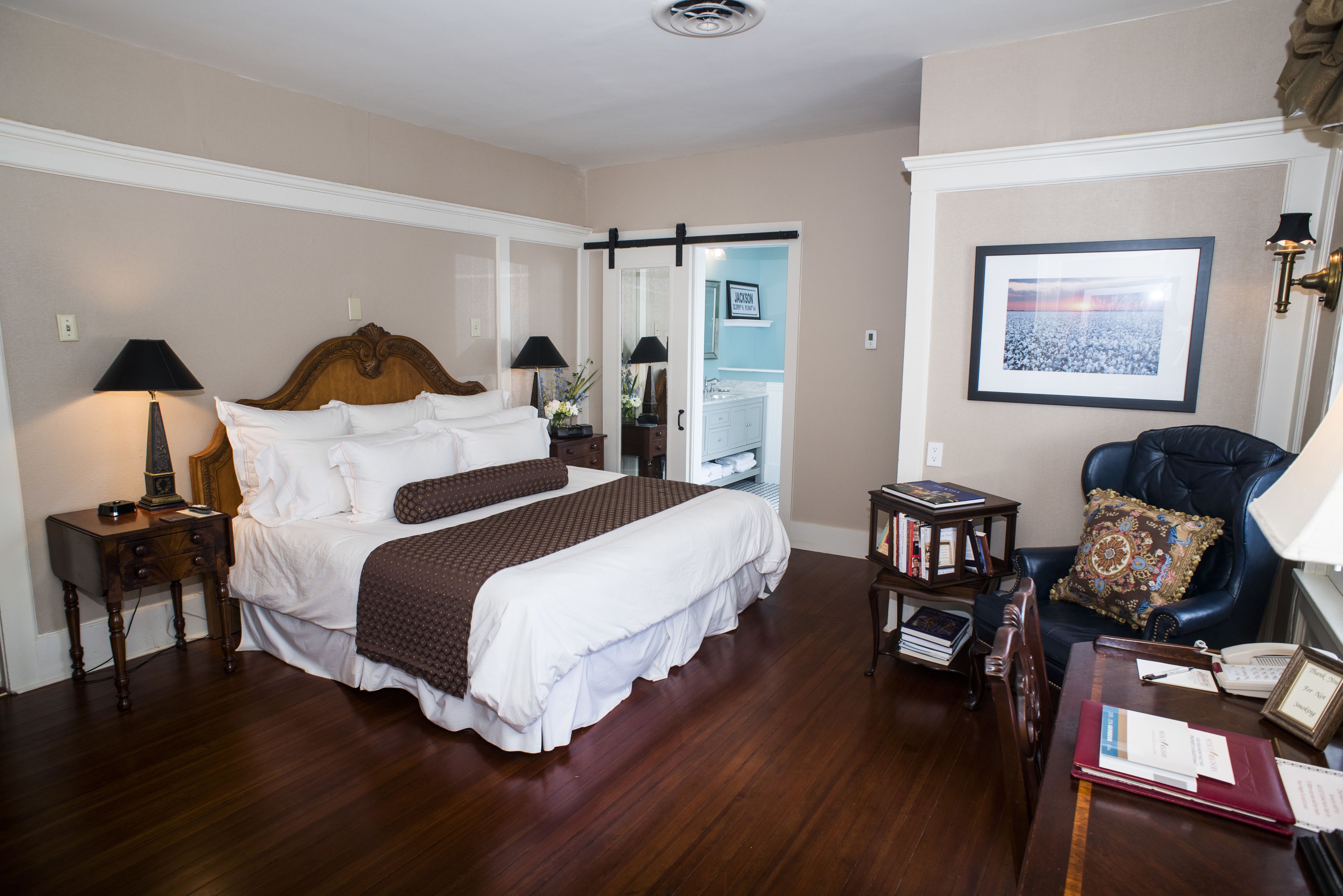 A hotel room with a large bed, two bedside tables with lamps, a chair with a decorative pillow, a small table with books and a phone, and a framed picture on the wall.