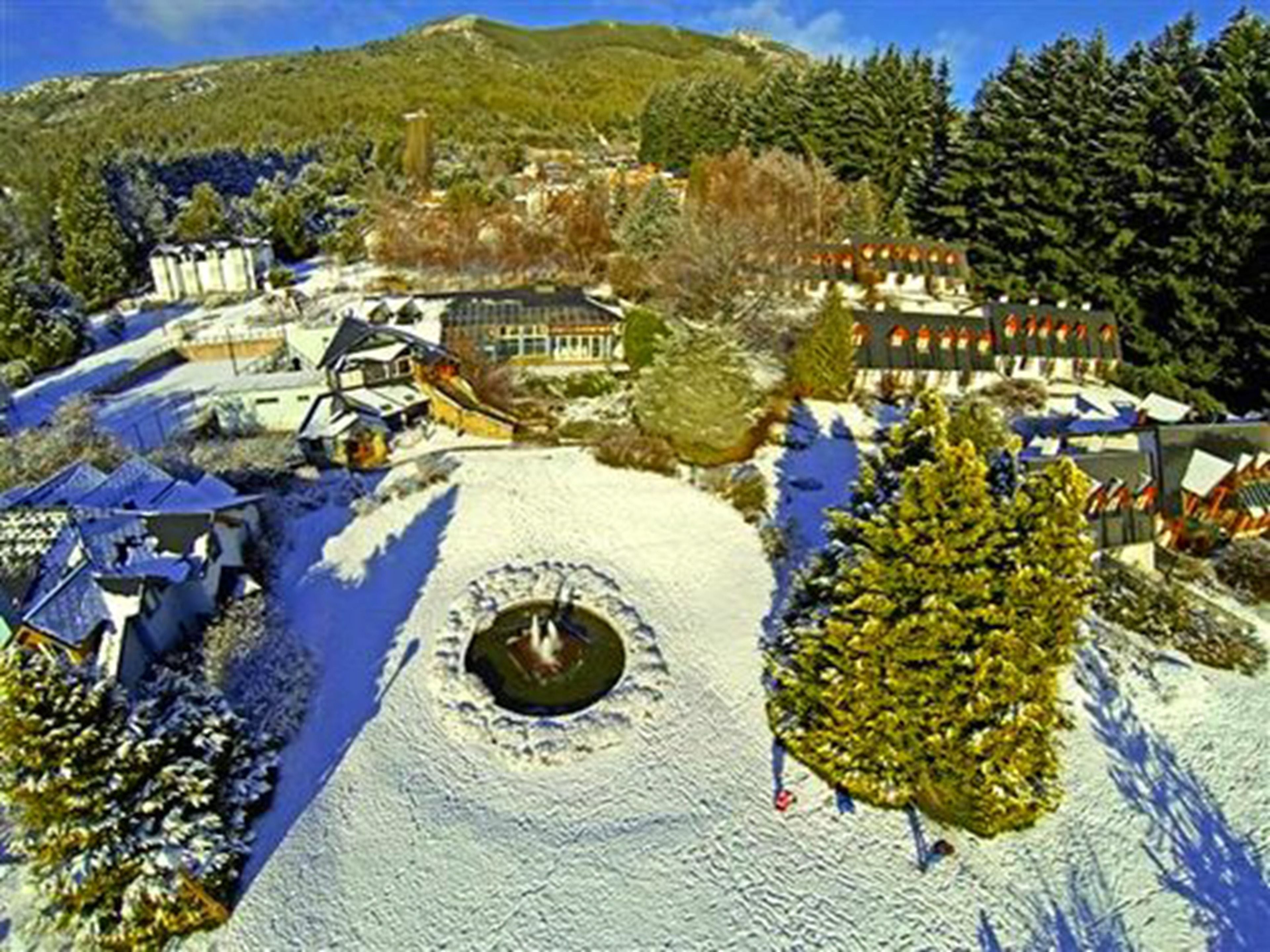 Paisagem coberta de neve com um edifício grande, uma estrutura circular e uma colina arborizada.