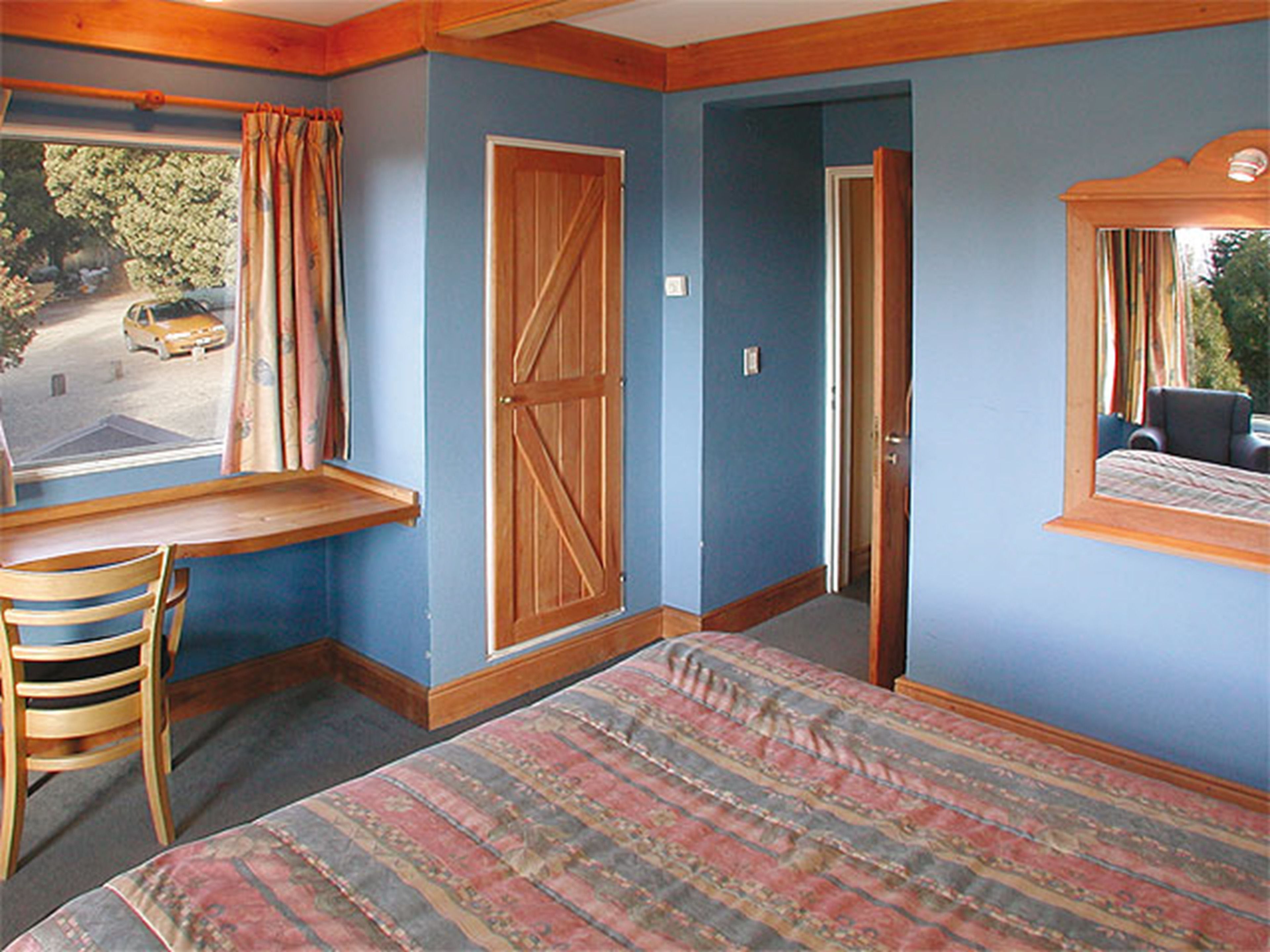 A room with a bed, a desk, and a chair. There is a window with curtains, a door, and a mirror on the wall.