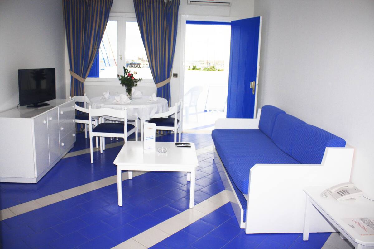 Ein Zimmer mit einem weißen Bett, einem weißen Schreibtisch, einem Fernseher, einem Esstisch mit Stühlen und einem Fenster mit Vorhängen.