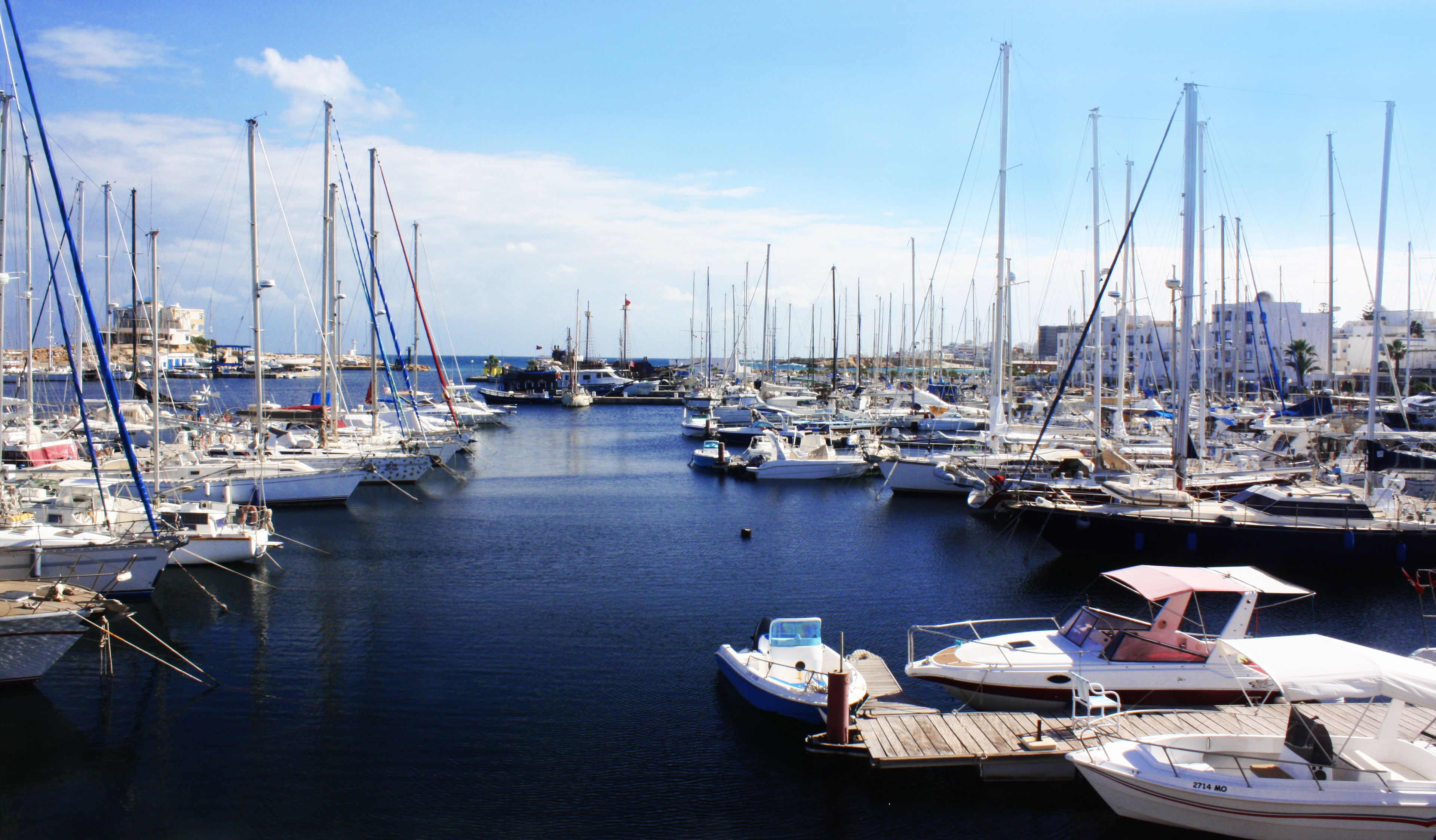 A marina with numerous boats docked, a clear sky, and buildings in the background.