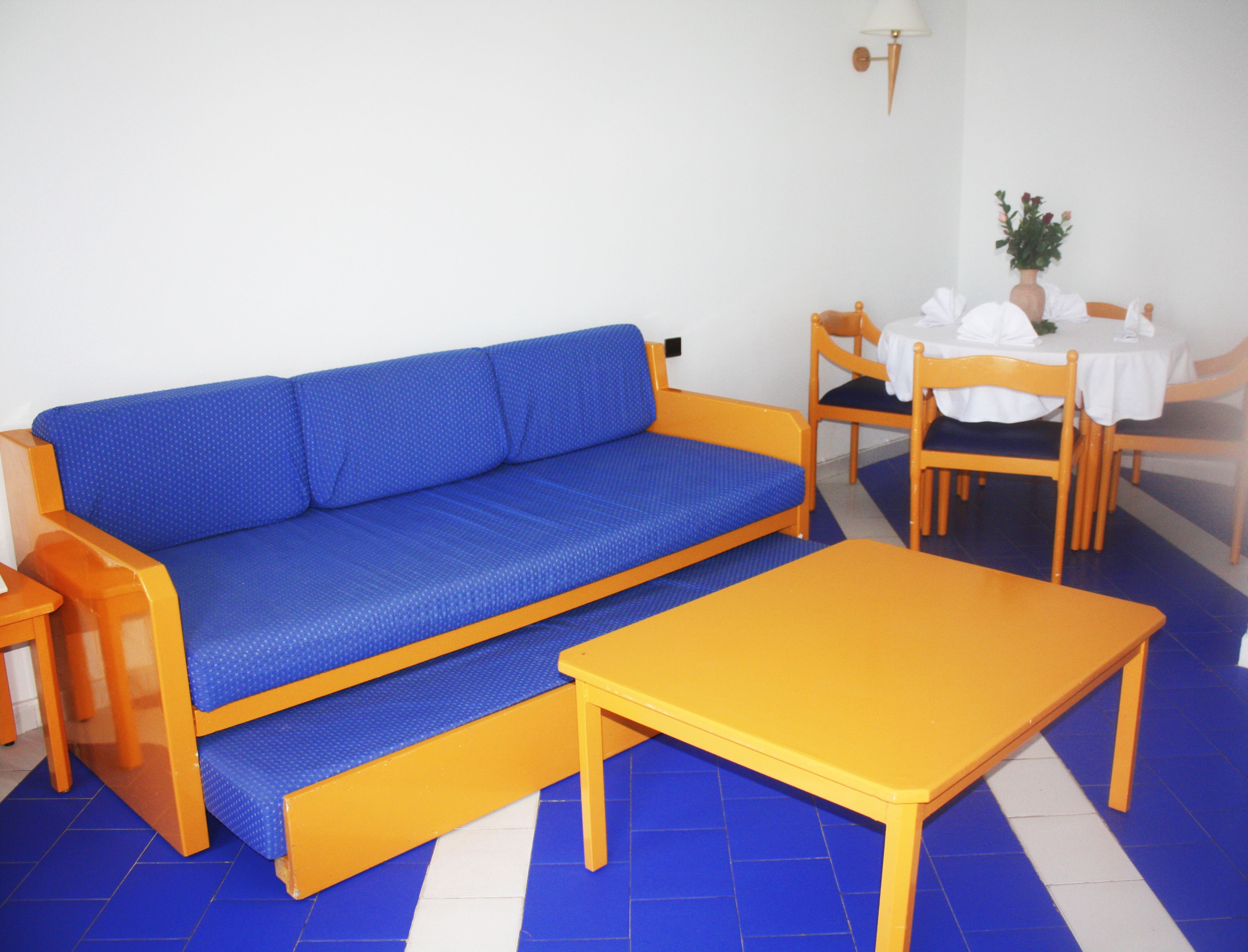 A room with a blue sofa, a wooden table, and a dining area with chairs and a table set for two.