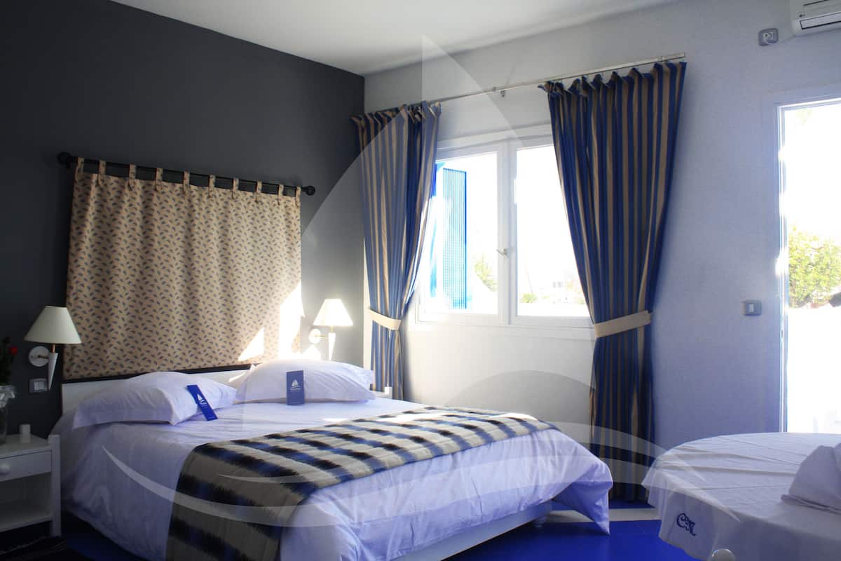 Ein Hotelzimmer mit einem Bett, einem Einzelbett, einem Fenster mit Vorhängen und einem Nachttisch mit Lampe.