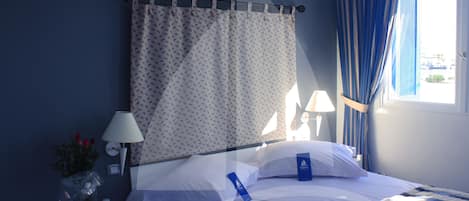 Una habitación de hotel con cama, mesita de noche con lámpara, jarrón con flores y ventana con cortinas.