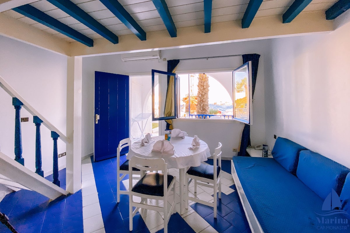 Ein Zimmer in Blau-Weiß gehalten, mit einem runden Tisch und Stühlen, einem Bett mit blauer Bettwäsche, einem Fenster mit Blick und einer Treppe mit blauen Geländern.