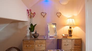 Un baño con tocador de vidrio, un espejo, una orquídea en maceta, un adorno con forma de corazón, una lámpara y un mantel con estampado.