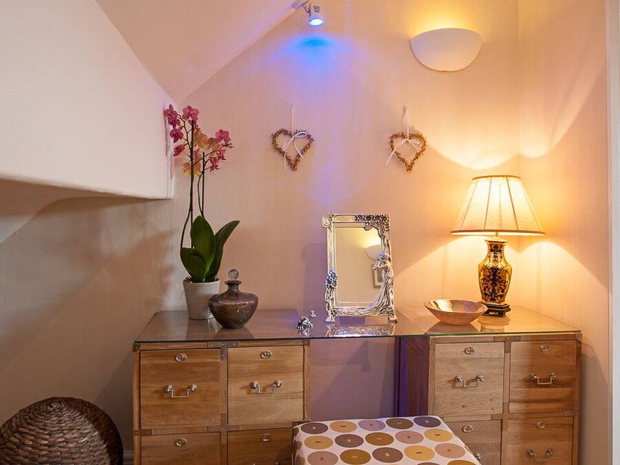 Un baño con tocador de vidrio, un espejo, una orquídea en maceta, un adorno con forma de corazón, una lámpara y un mantel con estampado.