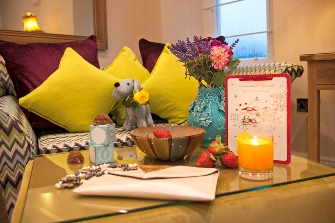 A cozy room with a sofa, pillows, a vase with flowers, a candle, and a framed card on a glass table.