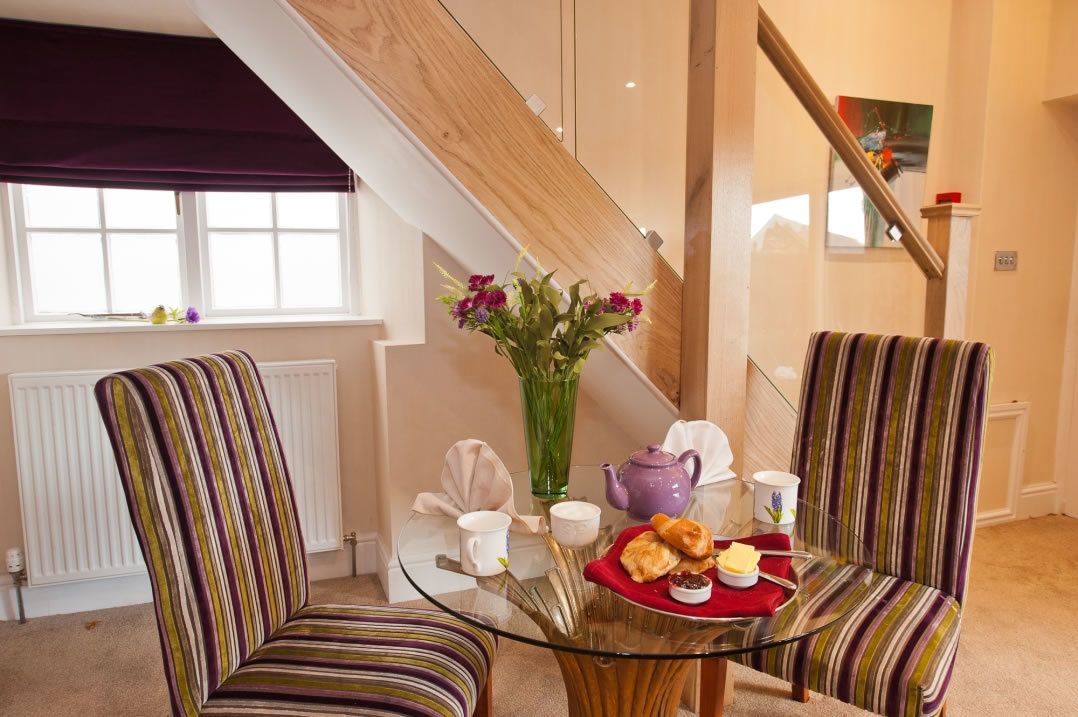 A cozy room with striped chairs, a glass table, a vase with flowers, and a tray of pastries.