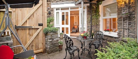 A patio with stone flooring, a wooden fence, and a table with chairs. There is a door leading to an interior space, and a window with plants on the right.