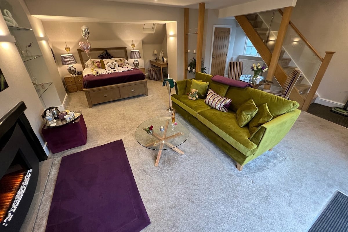 A modern living room with a green sofa, a glass coffee table, and a purple rug. There is a bed with a white headboard and a nightstand with a lamp. A staircase is visible in the background.