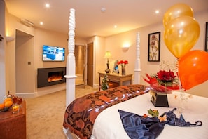 A hotel room with a bed, a fireplace, a television, and a desk with a lamp and flowers. - Windermere Boutique Hotel (Windermere)