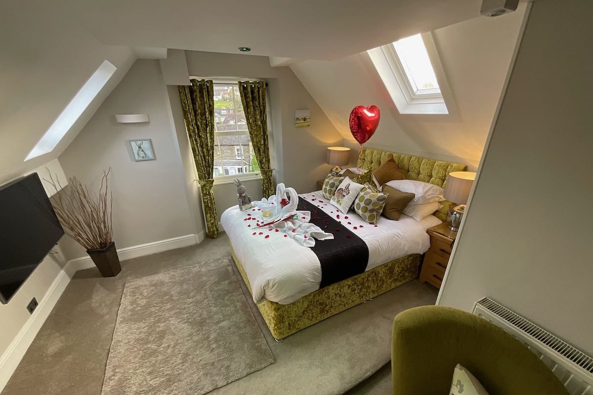 A bedroom with a bed, a bedside table, a lamp, a window with curtains, a plant, and a red heart-shaped balloon.