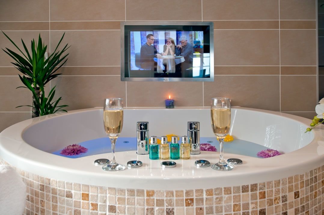 A bathtub with two champagne glasses, a television mounted above it, and a small table with toiletries.