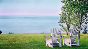 Two Adirondack chairs facing a body of water with a grassy foreground and a tree to the right. - Auberge de la Baie (Caraquet)