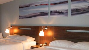 A hotel room with two beds, a wooden headboard, bedside lamps, and a wall-mounted artwork. - Auberge de la Baie (Caraquet)