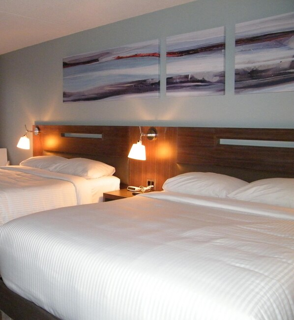 Habitación de hotel con dos camas, cabecero de madera, lámparas de noche y una obra de arte colgada en la pared.