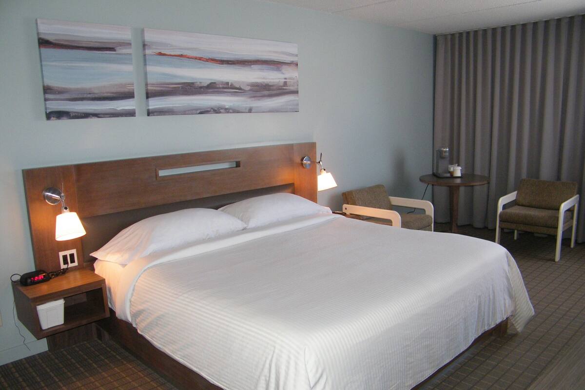 Een hotelkamer met een groot bed, twee bedlampen, een nachtkastje, een stoel, een kleine tafel en twee abstracte schilderijen aan de muur.