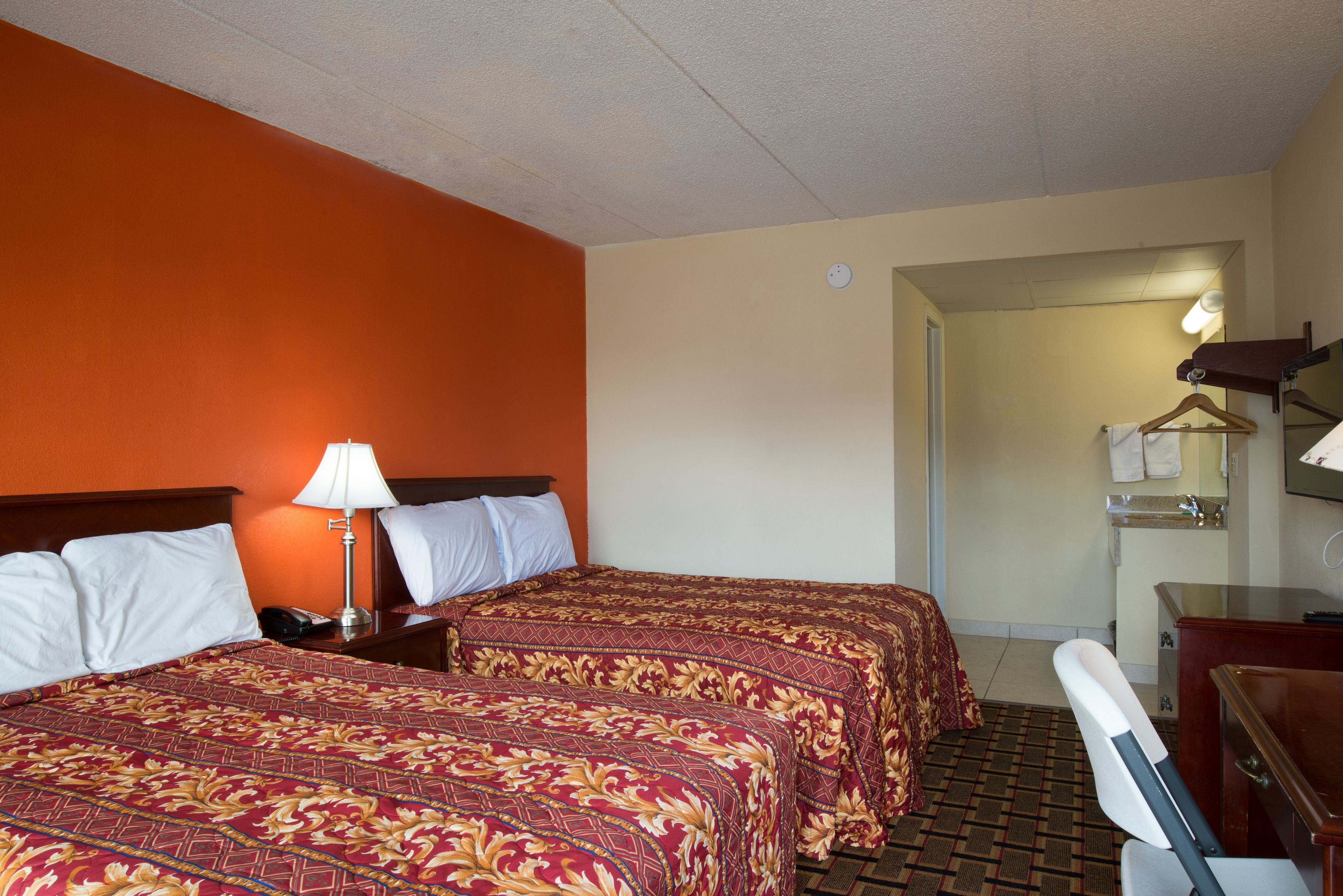 Quarto de hotel com duas camas, uma escrivaninha, uma cadeira, um abajur e um banheiro visível por uma porta aberta.