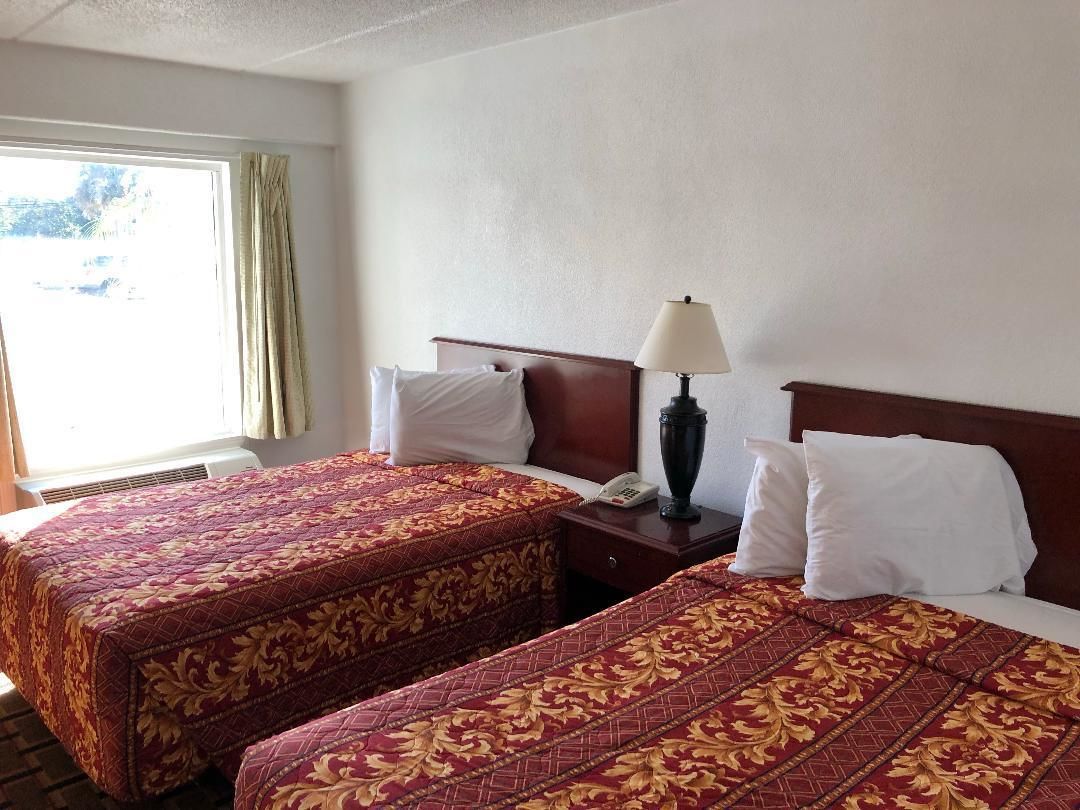 A hotel room with two beds, each with a red and gold patterned bedspread, a bedside table with a lamp, and a window with curtains.