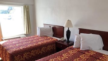 A hotel room with two beds, each with a red and gold patterned bedspread, a bedside table with a lamp, and a window with curtains.
