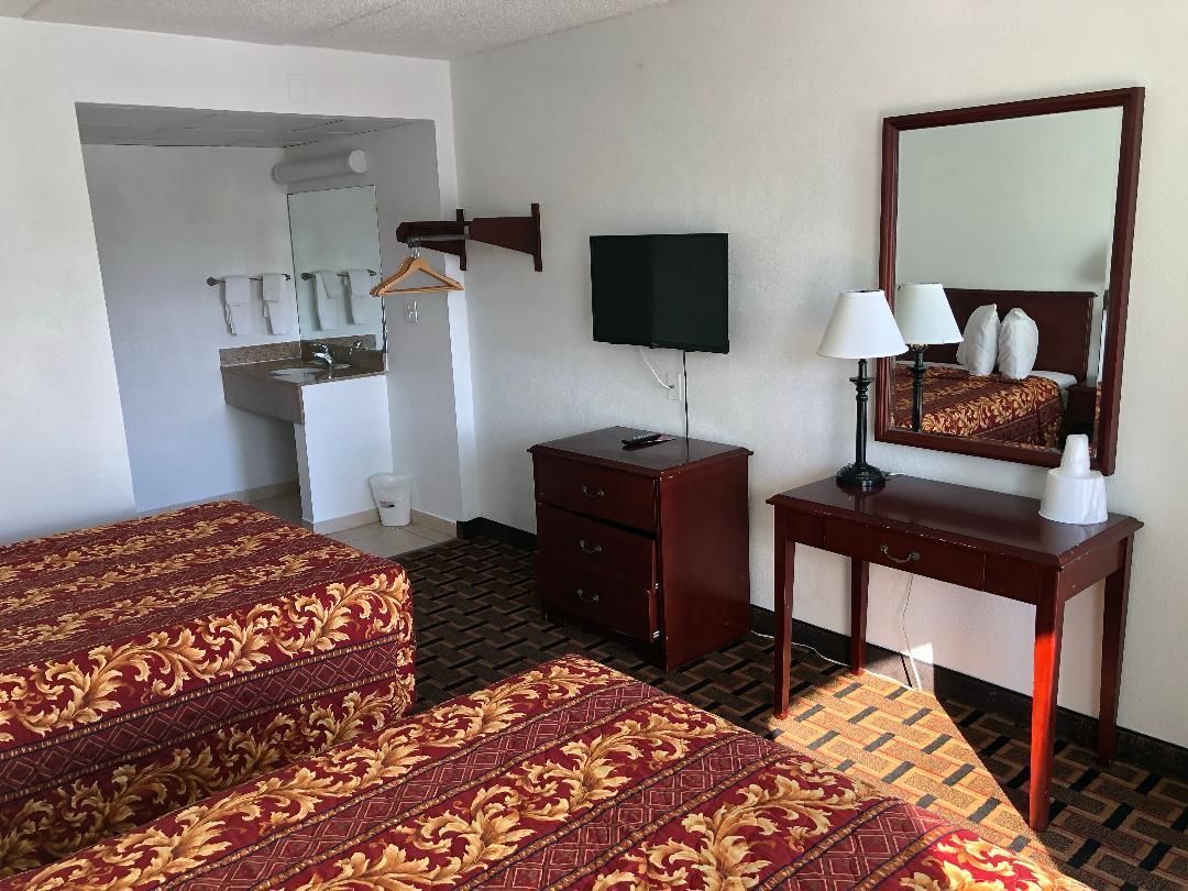 Habitación de hotel con dos camas, televisor, escritorio con lámpara, espejo y baño con lavamanos y toallero.