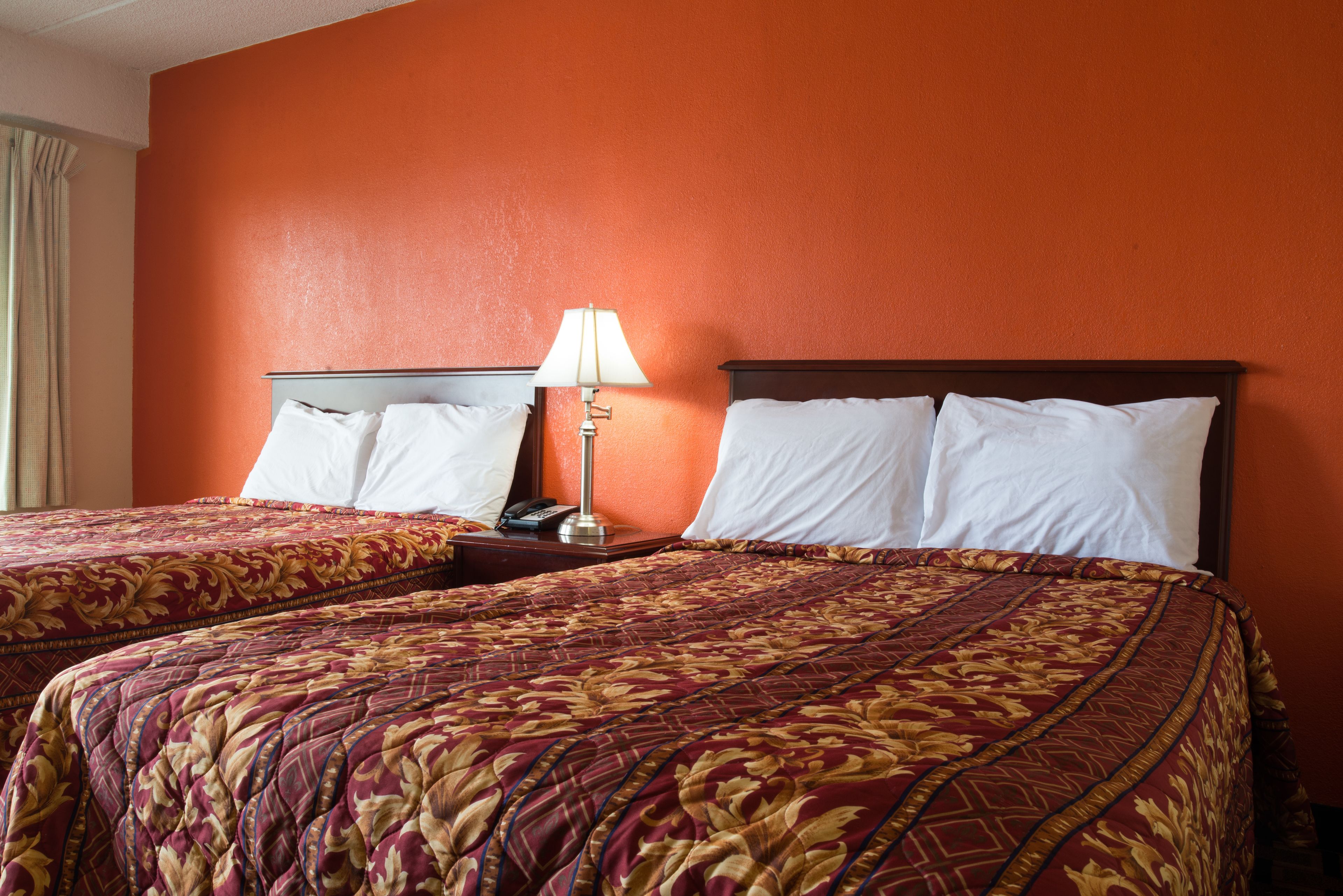 Habitación de hotel con dos camas, cada una con una colcha de diseño rojo y dorado, una mesita de noche con lámpara y una pared naranja.