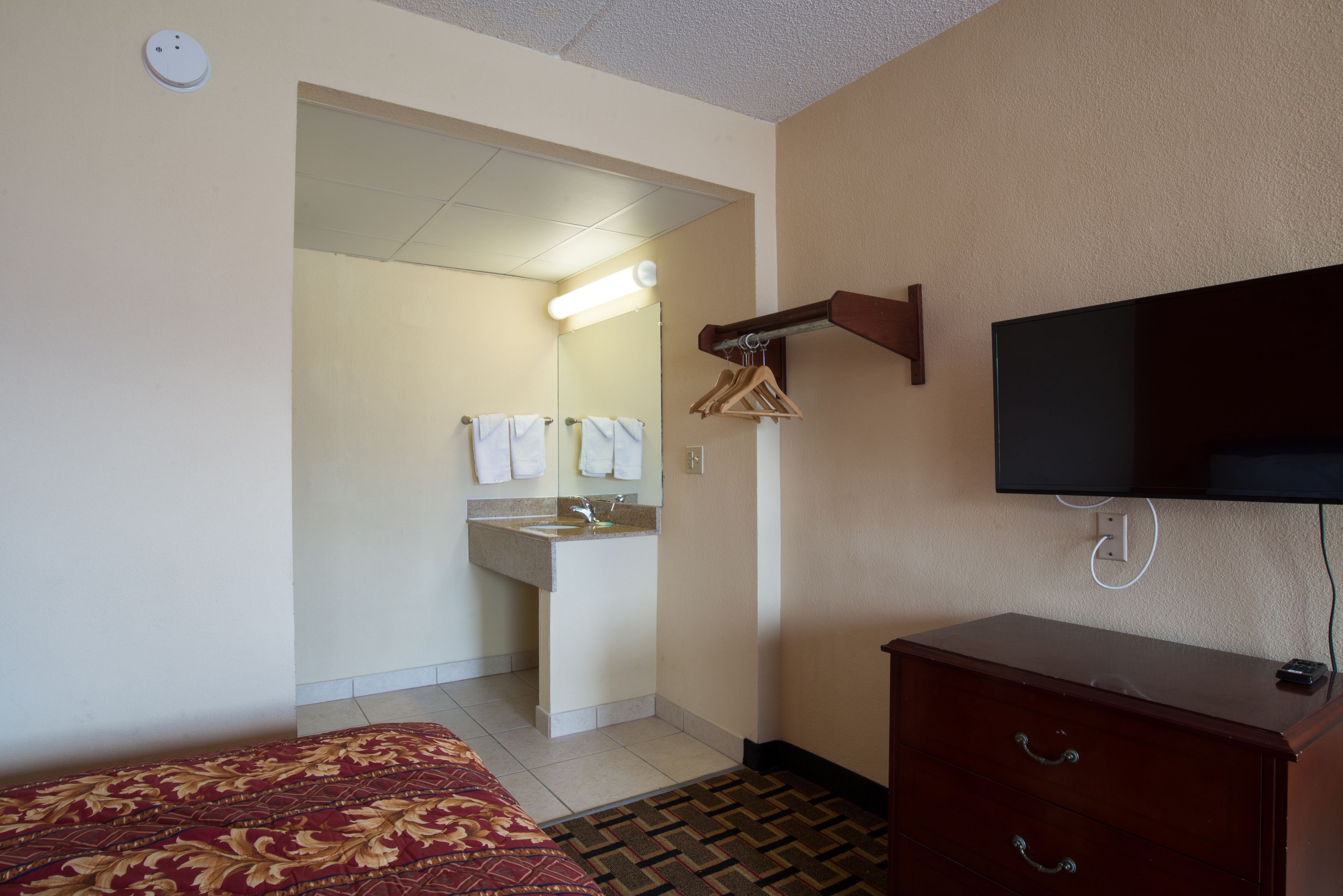 A hotel room with a bed, a dresser, a television, a bathroom with a sink and mirror, and a hallway leading to another room.