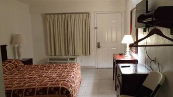 Una habitación de hotel con cama, escritorio, silla, lámpara, perchero y puerta.
