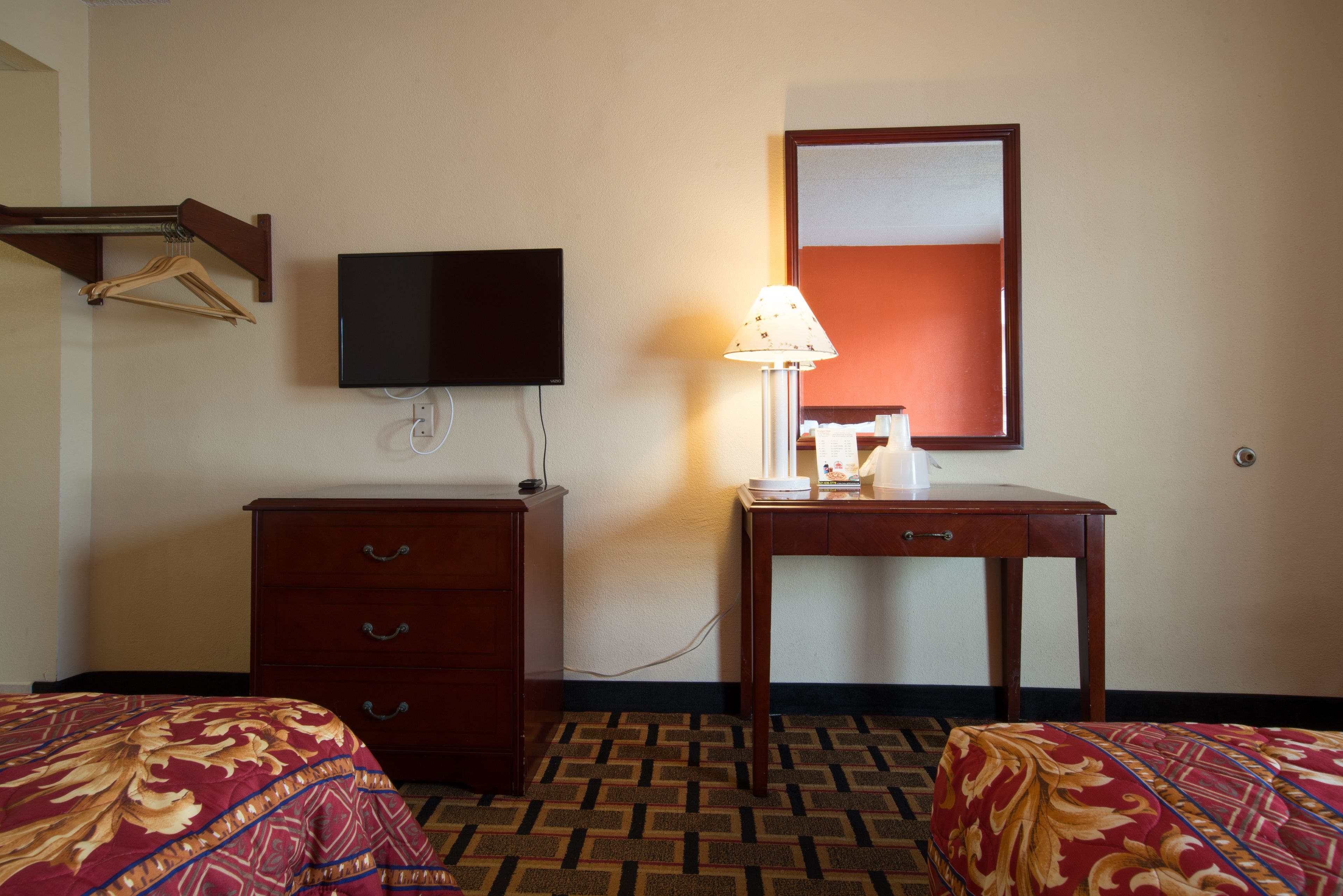 A hotel room with a bed, a dresser, a desk with a lamp, a television, and a mirror.