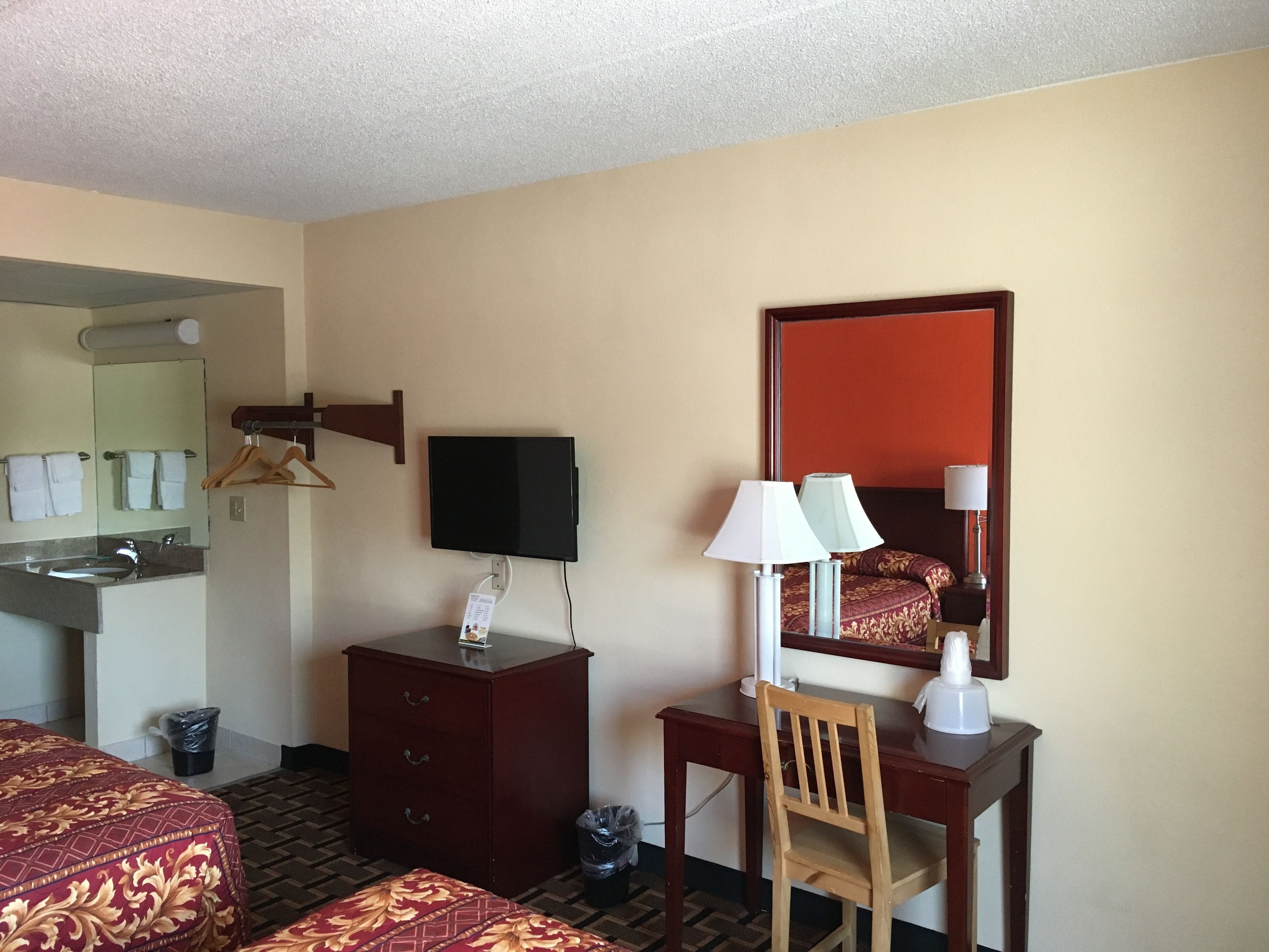 Ein Hotelzimmer mit Bett, Schreibtisch mit Stuhl, Fernseher, Spiegel, Kommode und einem Badezimmer mit Waschbecken und Handtuchhalter.