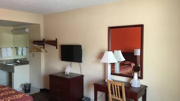 Habitación de hotel con cama, escritorio con silla, televisión, espejo, cómoda y baño con lavabo y toallero.