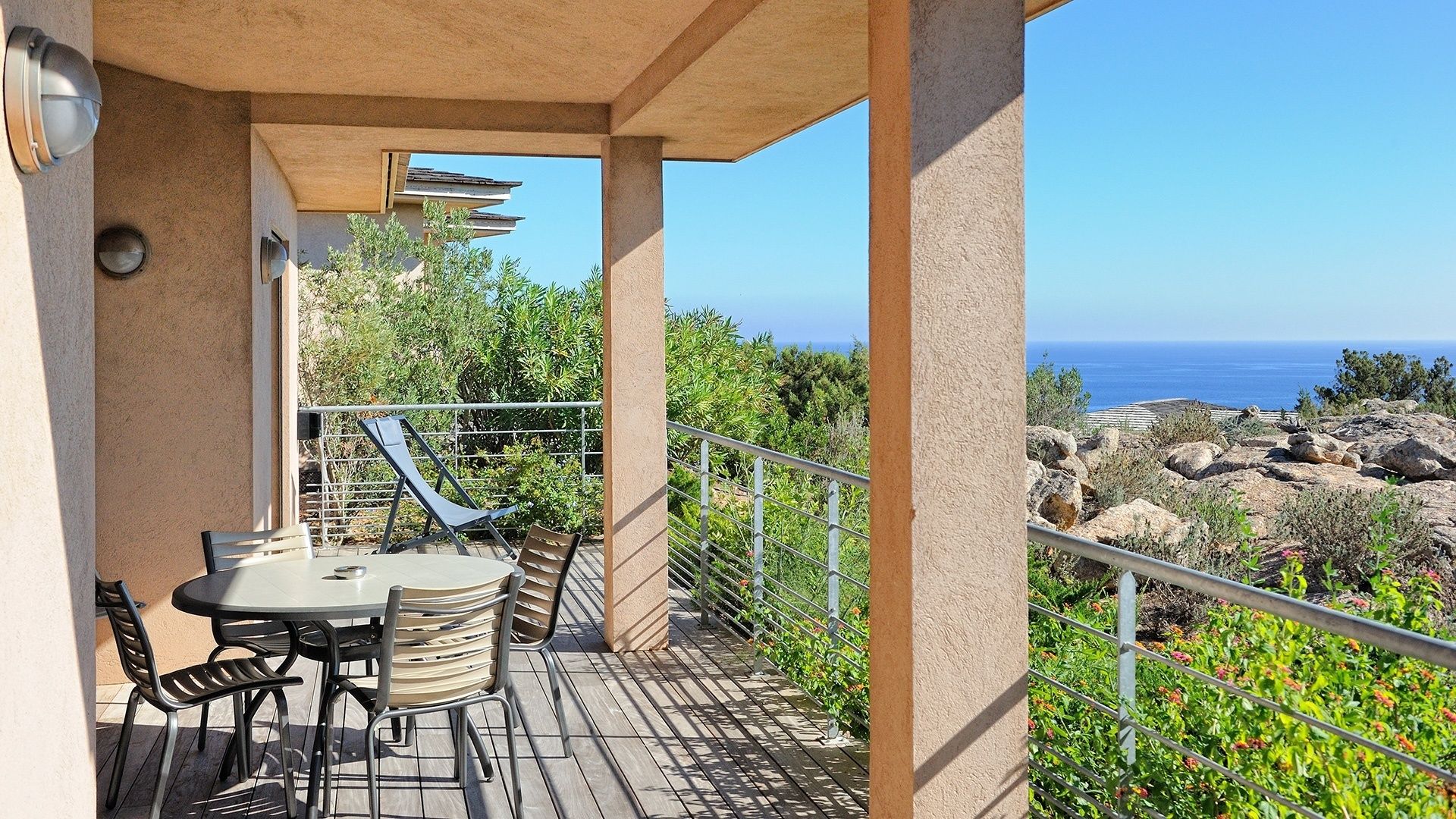 A balcony with a table and chairs, overlooking a rocky landscape and the sea.