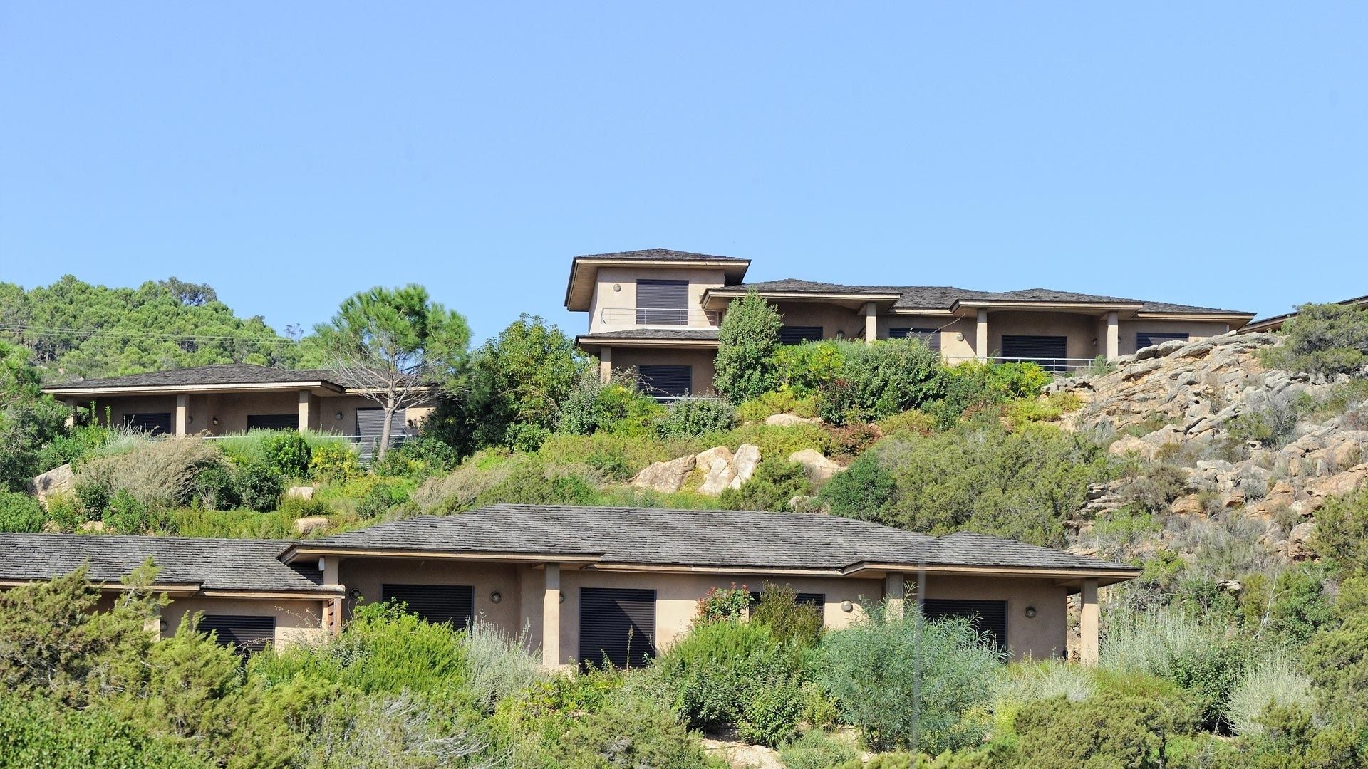 A hillside residential area with multiple houses featuring similar architectural styles, surrounded by natural vegetation and rocky terrain.
