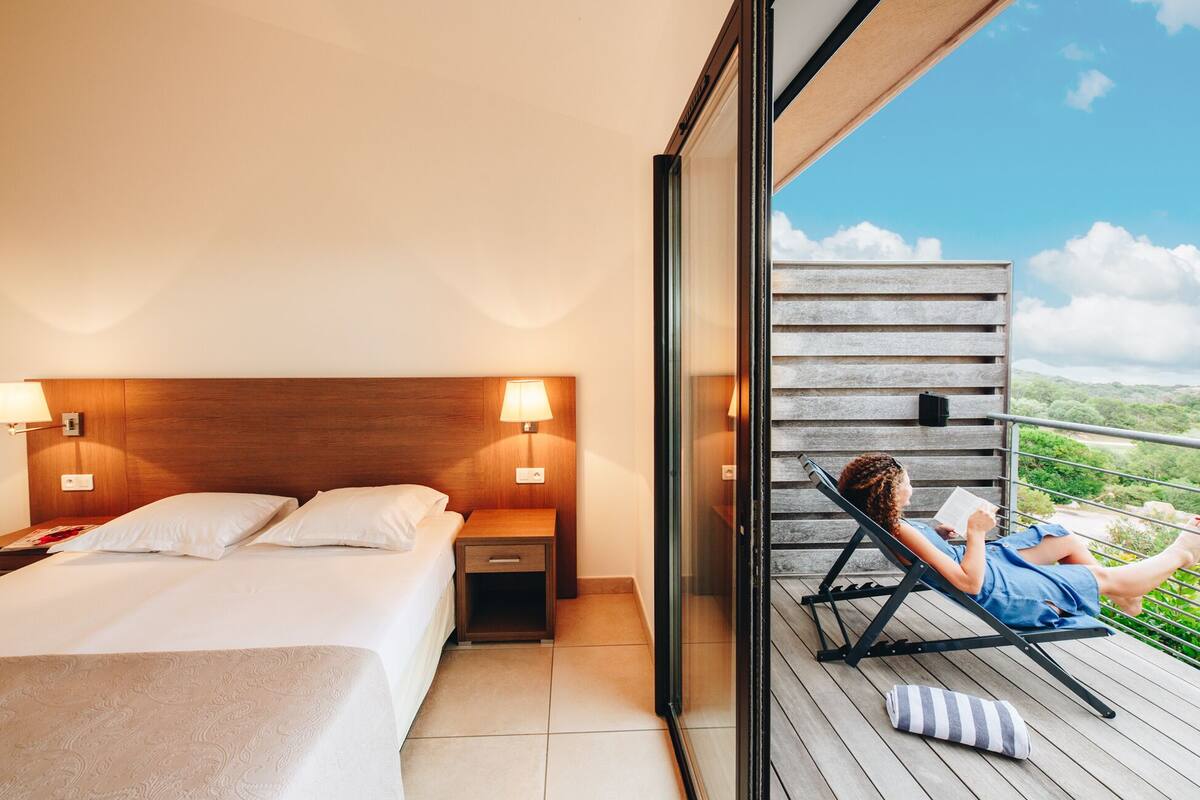 A hotel room with a bed, bedside lamps, a nightstand, and a balcony with a view of the landscape.