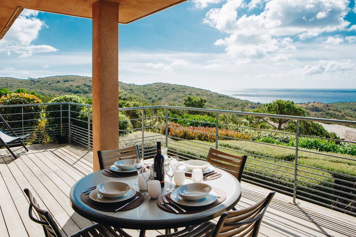 A round table set for four with plates, glasses, and cutlery, overlooking a scenic view of hills and the sea.