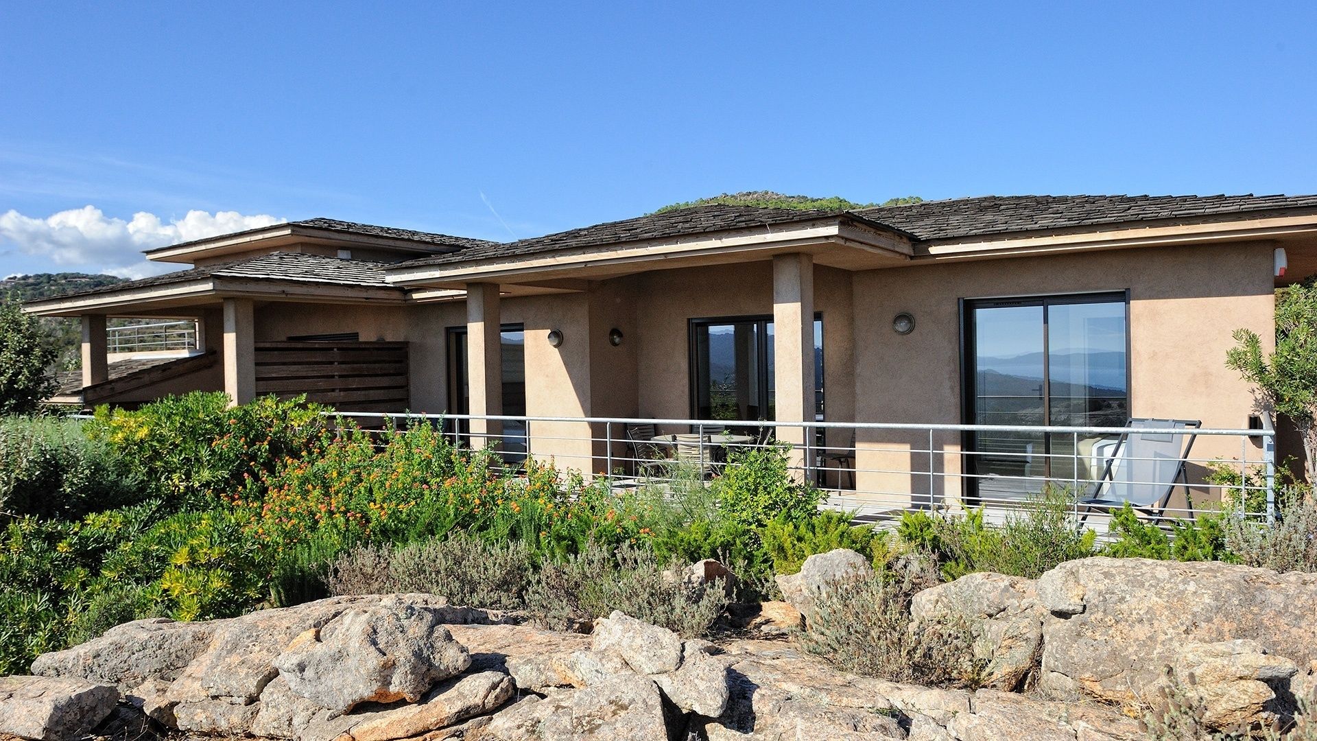 A modern two-story house with a balcony, large windows, and a view of the surrounding landscape.