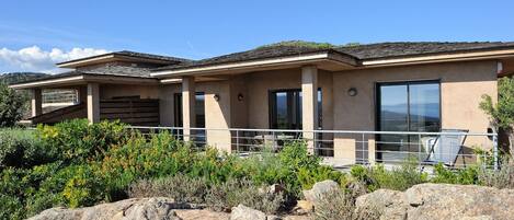 A modern two-story house with a balcony, large windows, and a view of the surrounding landscape.