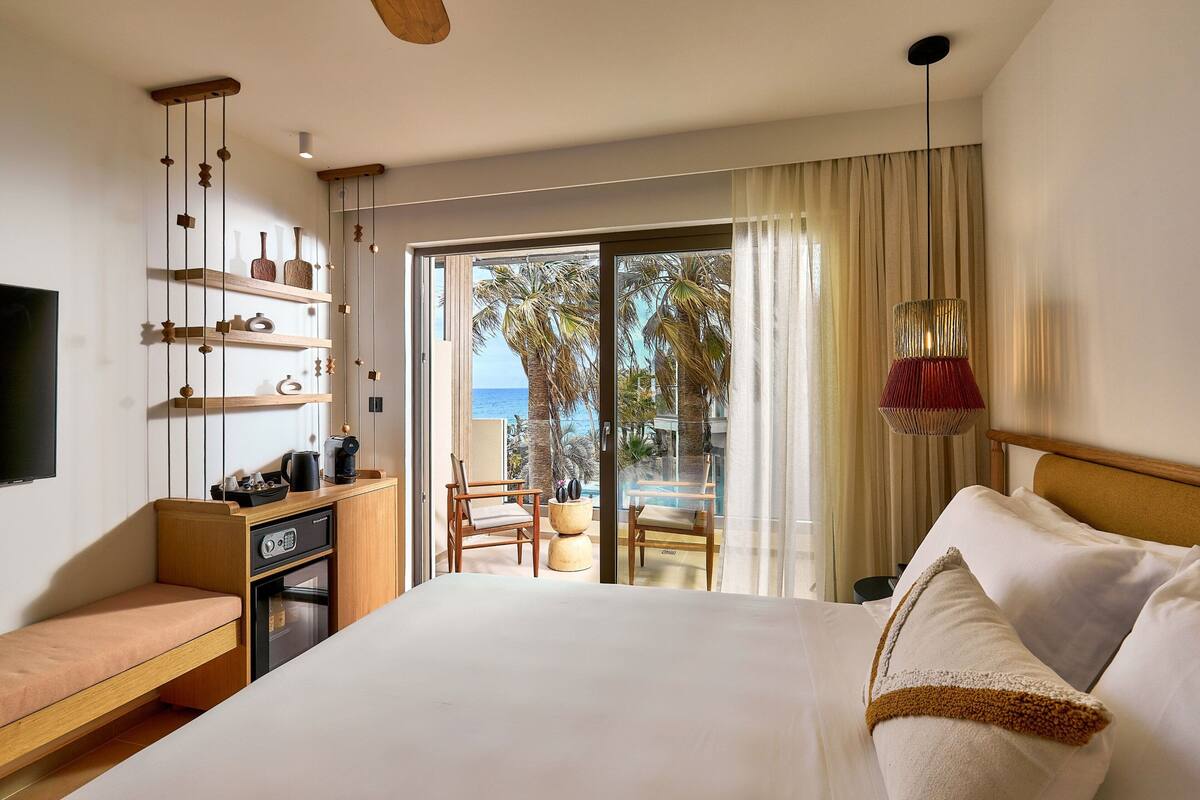 A hotel room with a large bed, a wooden bench, a small kitchenette, and a balcony with a view of palm trees and the sea.