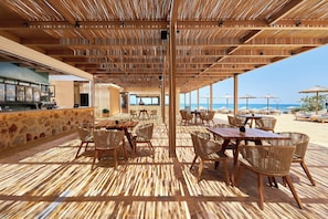 An outdoor dining area with wooden tables and chairs, a bar area, and a view of the beach. - Alai, Crete, a Tribute Portfolio Resort (Hersonissos)