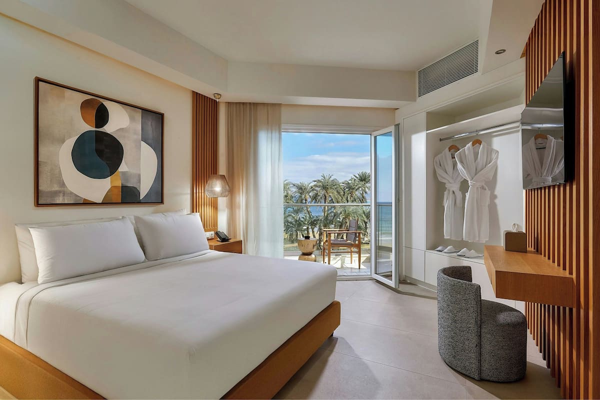 A modern hotel room with a large bed, a balcony with a view of palm trees, and a wooden vanity with a mirror.