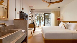 Habitación de hotel con una cama grande, ventilador de techo de madera, baño con lavamanos amplio y vista a palmeras a través de la ventana.