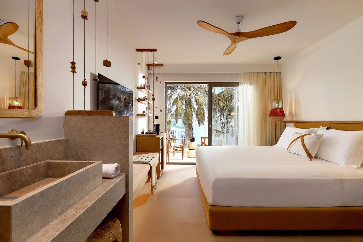 A hotel room with a large bed, a wooden ceiling fan, a bathroom with a large sink, and a view of palm trees through the window.