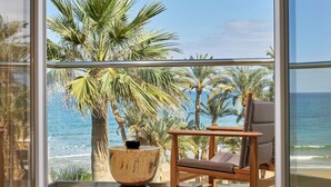 Un balcón con una silla de madera y una mesa redonda, con vista a una playa con palmeras y el océano.