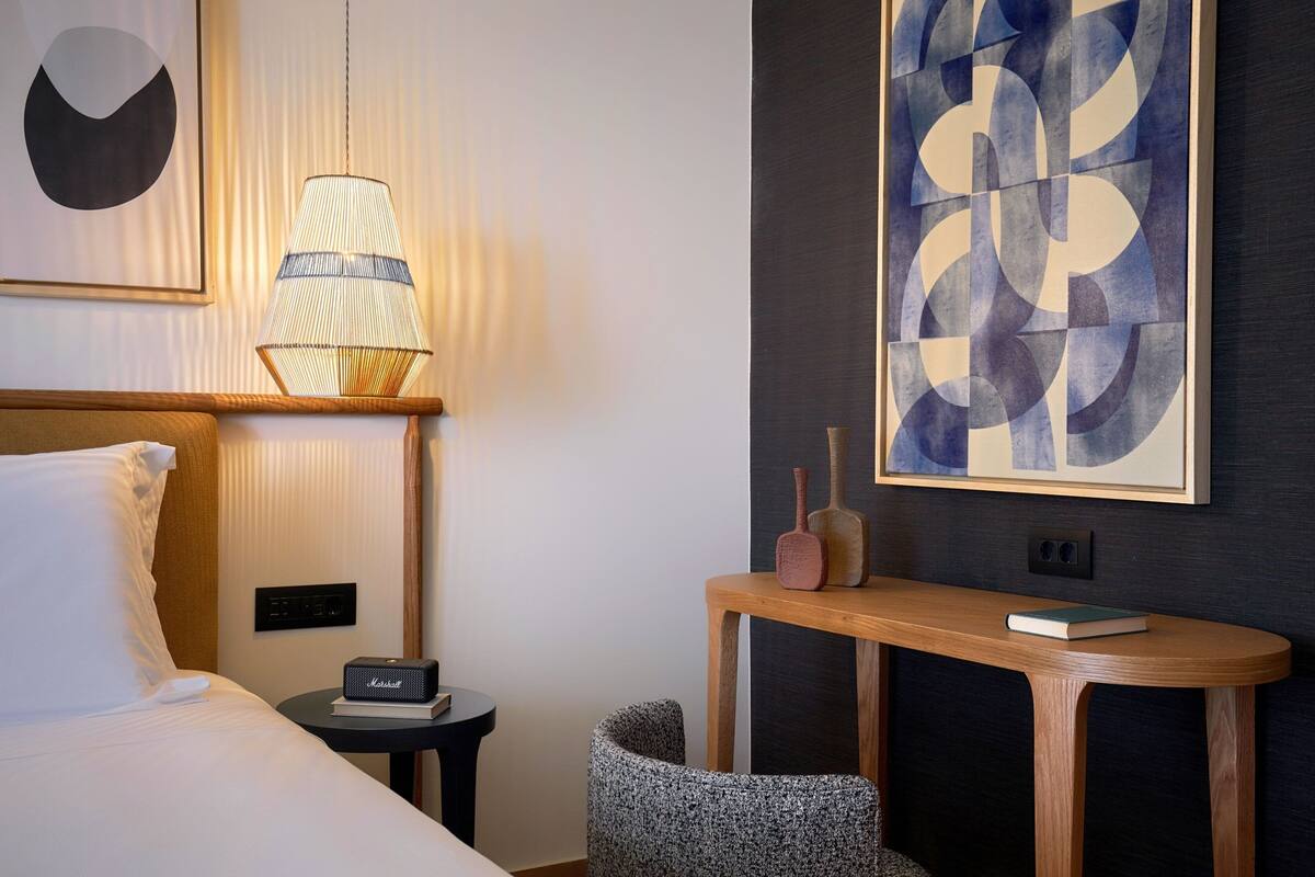A hotel room with a bed, a lamp, a bedside table with a book, a small chair, and artwork on the walls.