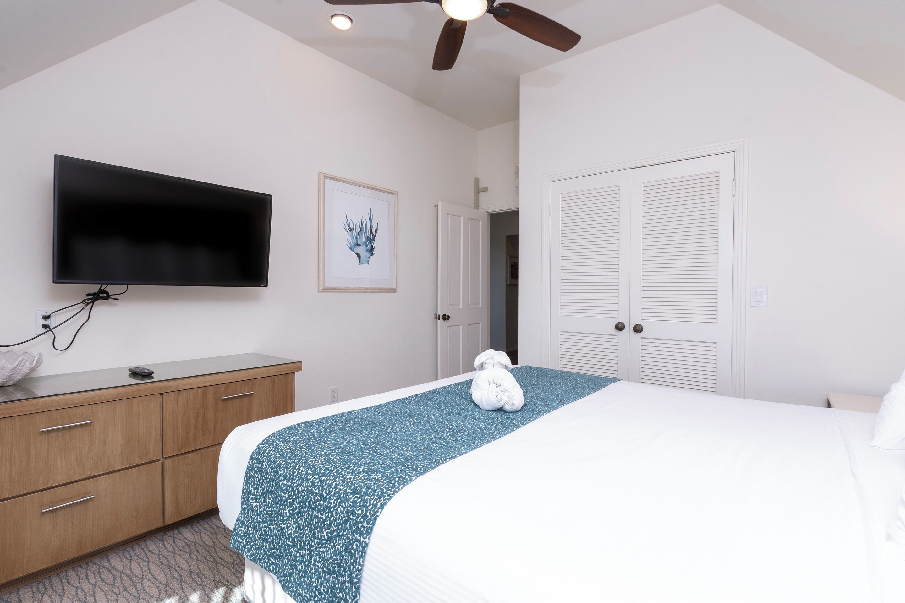 Une chambre d’hôtel avec un lit, un téléviseur fixé au mur, une commode en bois et un ventilateur de plafond.
