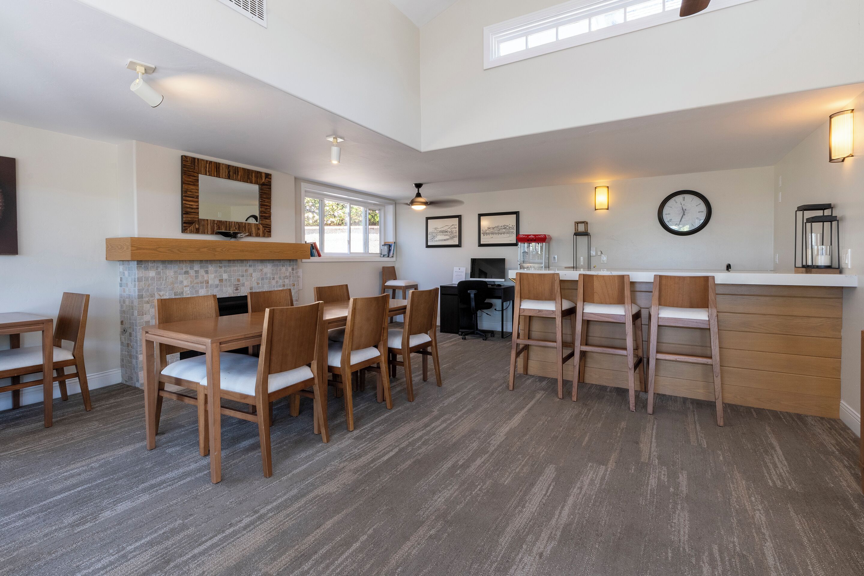 A spacious dining area with wooden tables and chairs, a kitchen in the background, and a clock on the wall.