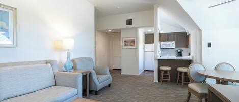 A hotel room with a sofa, a small table, a lamp, a kitchenette, and a dining area with chairs.