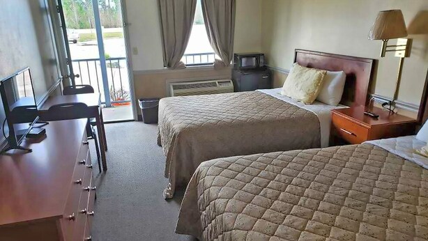 A hotel room with two beds, a desk with a chair, a lamp, and a view of the outdoors. - Traveler's Choice Motel (Bay St Louis)