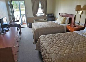 A hotel room with two beds, a desk with a chair, a lamp, and a view of the outdoors. - Traveler's Choice Motel (Bay St Louis)